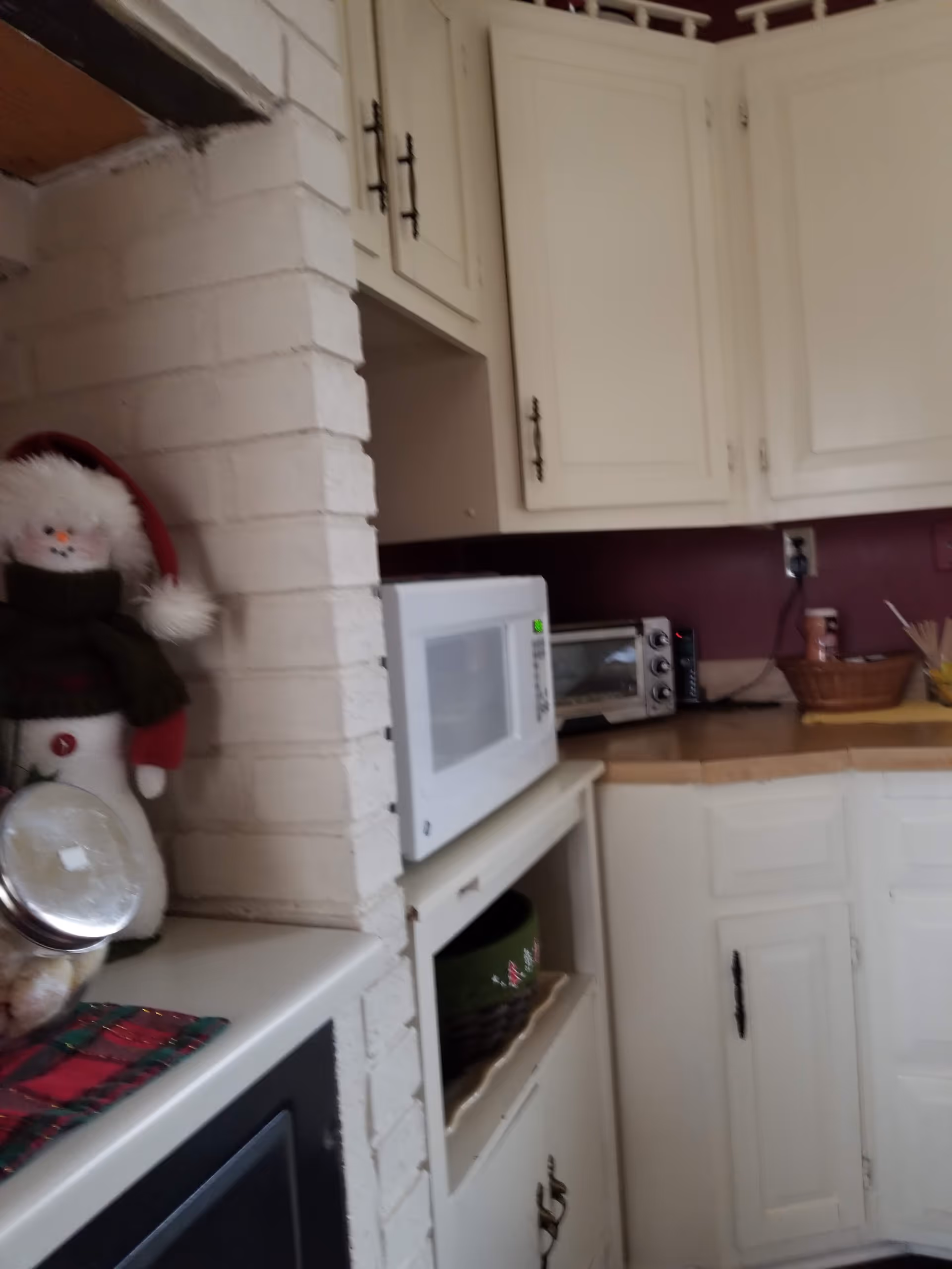 White kitchen with cabinets, microwave, toaster oven, brick pillar, and holiday snowman decoration.