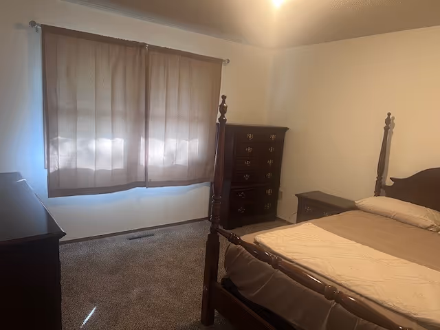 Dark wood bedroom with four-poster bed, dresser, nightstand, and window with brown curtains.
