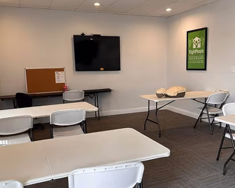 Classroom-style room with white tables, folding chairs, a wall-mounted TV, and CPR mannequins.