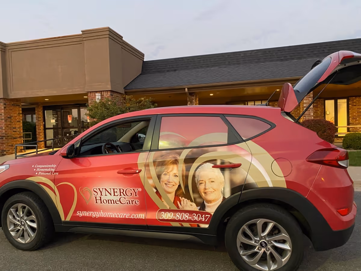 A pink Synergy HomeCare van parked in front of a brick facility entrance.