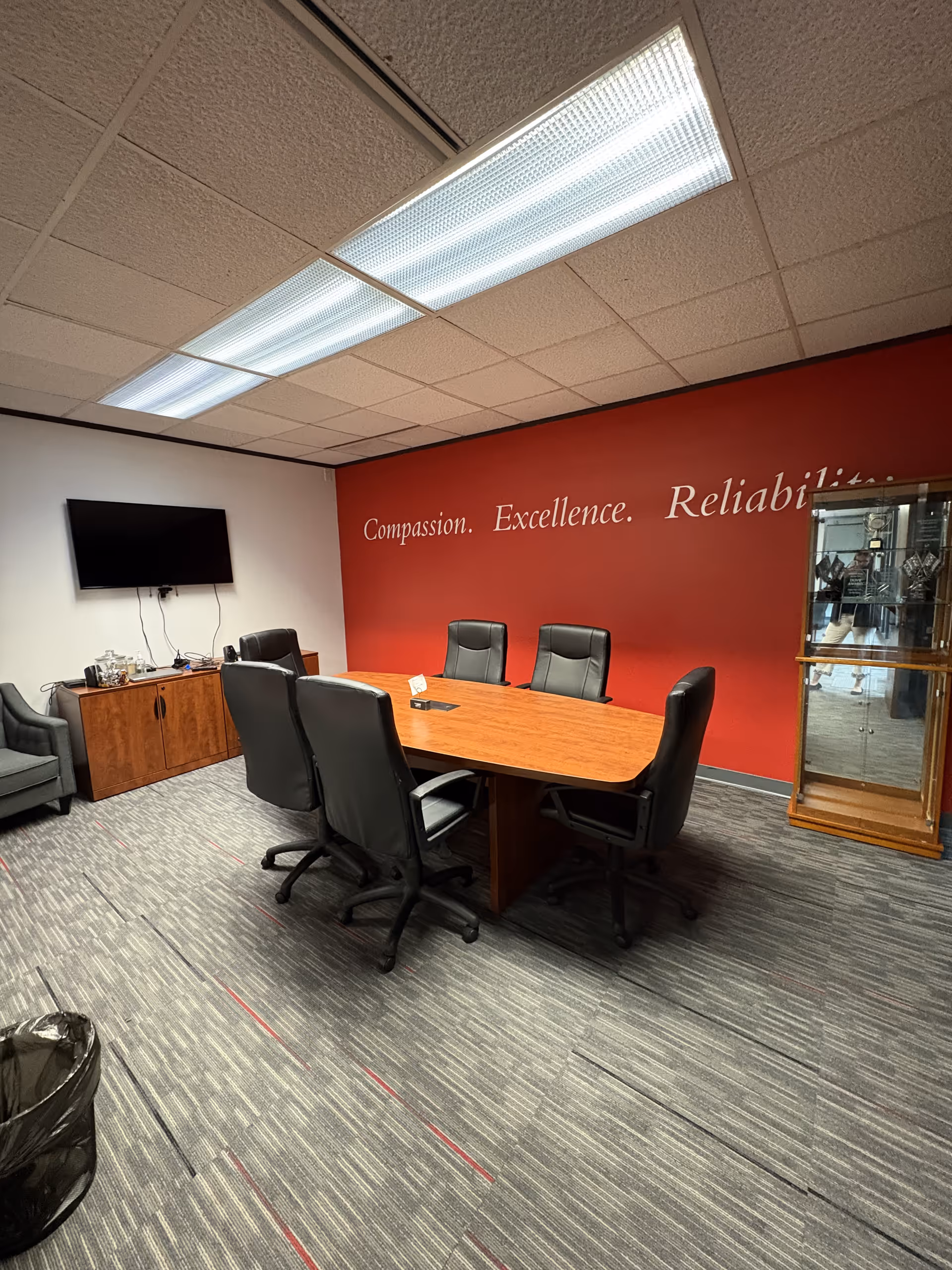 A conference room with a wooden table, black chairs, TV, and a red wall slogan.
