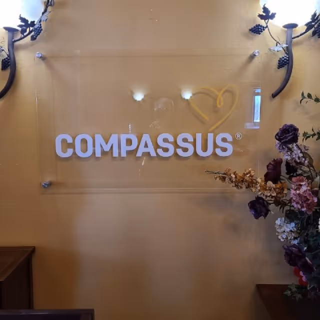 COMPASSUS sign mounted on a beige wall with decorative flowers nearby.