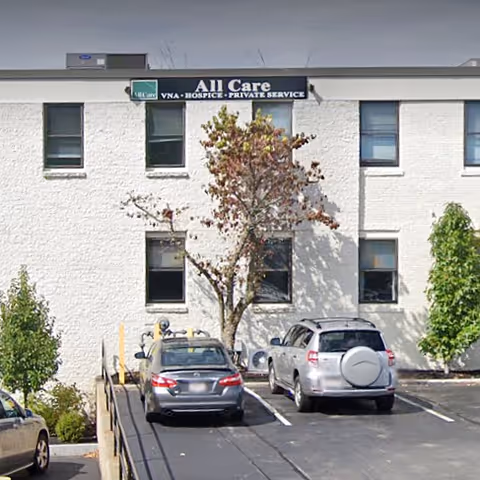 Exterior view of All Care facility with sign, parked cars, and trees.