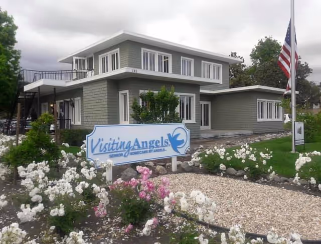 Two-story green-gray senior care building with landscaped front garden, flag, and Visiting Angels sign.