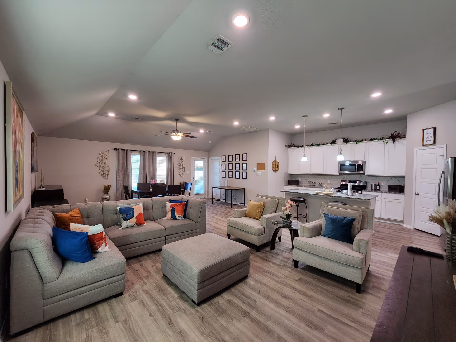 Spacious open-concept living area with grey sofas, ottoman, and kitchen in back.