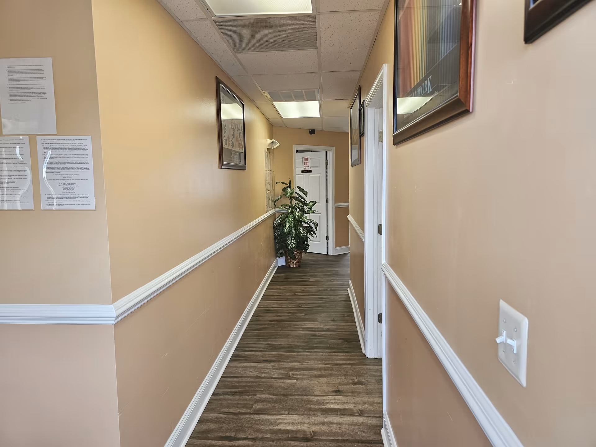 Long beige hallway in a senior facility with framed artwork and plant at the end.