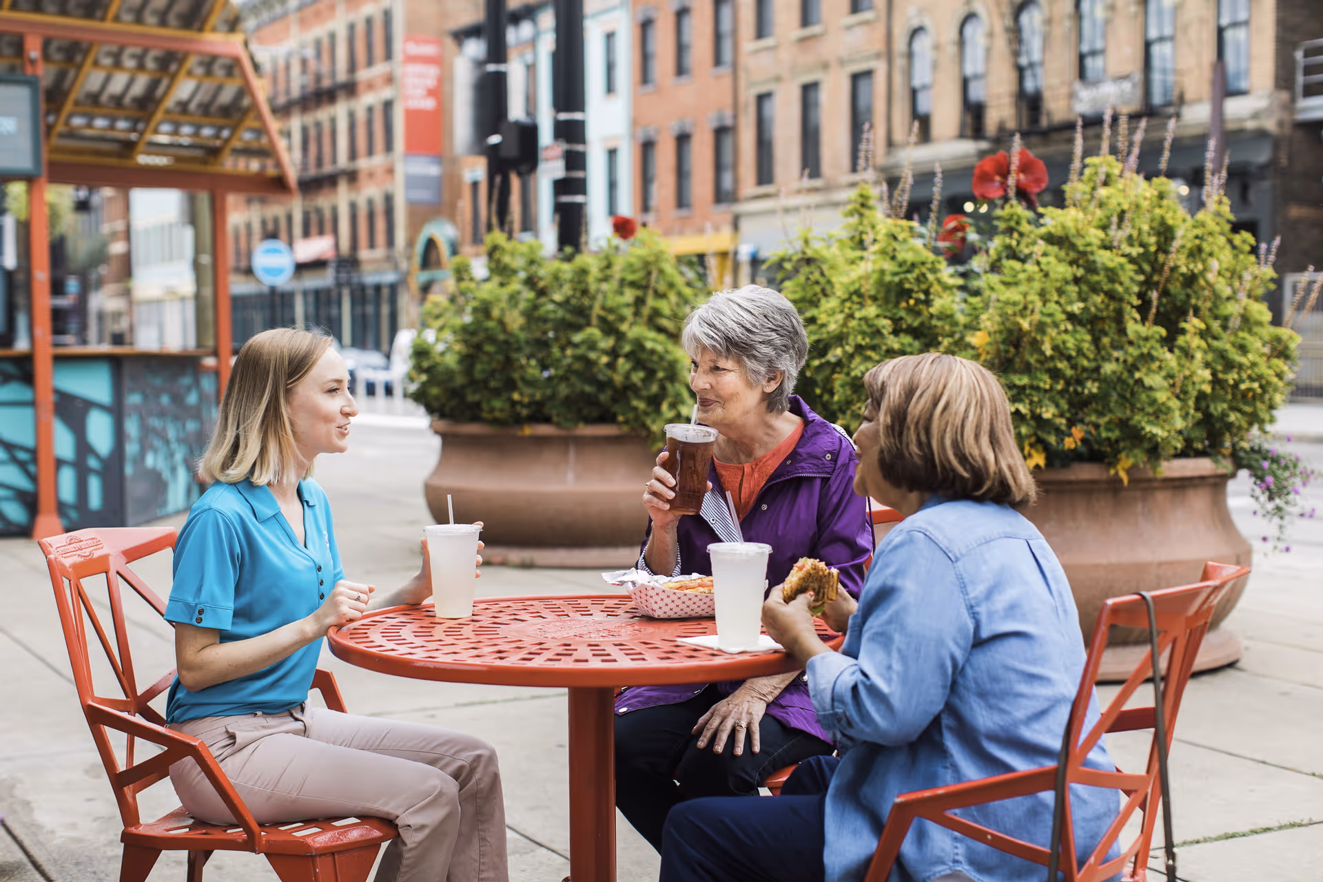 Three people sit at a red metal table outdoors, enjoying drinks and snacks.