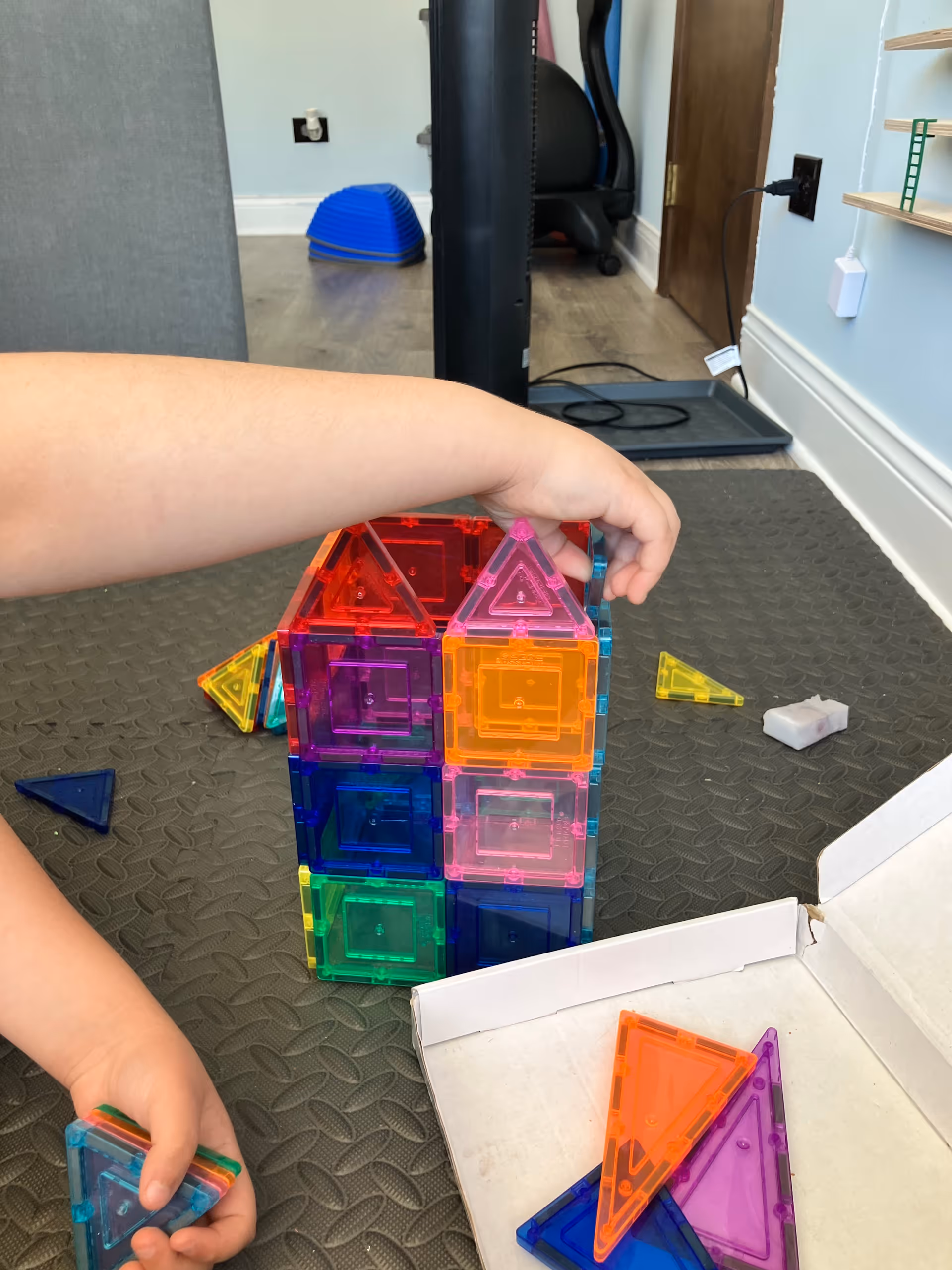 A person stacks colorful translucent magnetic tiles into a tower on a black play mat.