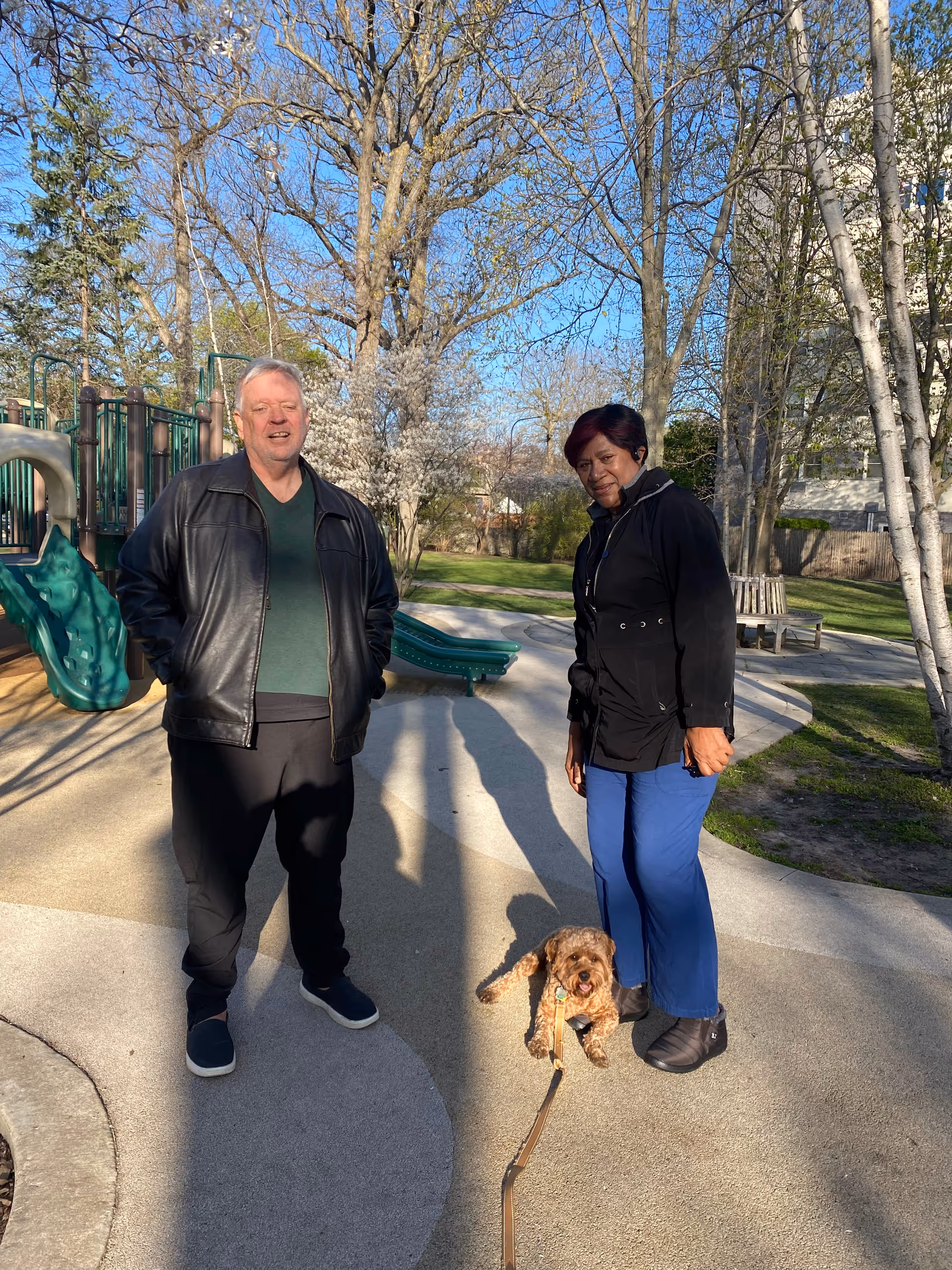 Two adults and a small dog stand on a paved outdoor park area near playground.