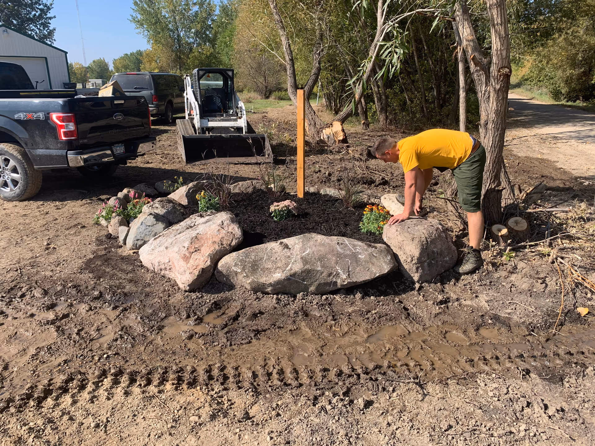 Person in a yellow shirt arranges large rocks around a garden bed outdoors.