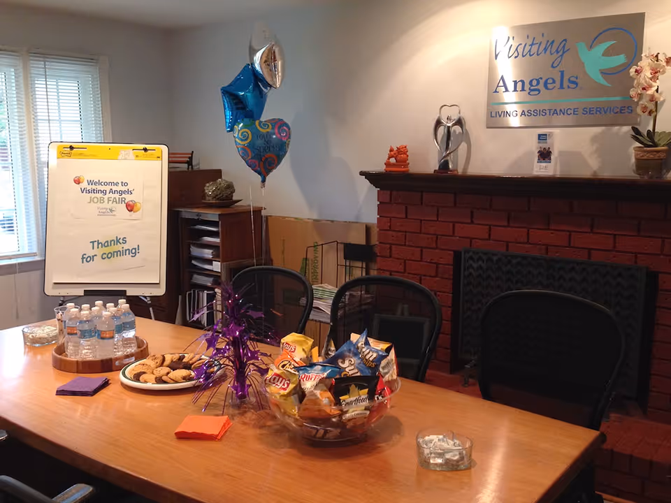 Indoor meeting room with a snack table, balloons, and a Visiting Angels sign on the wall.