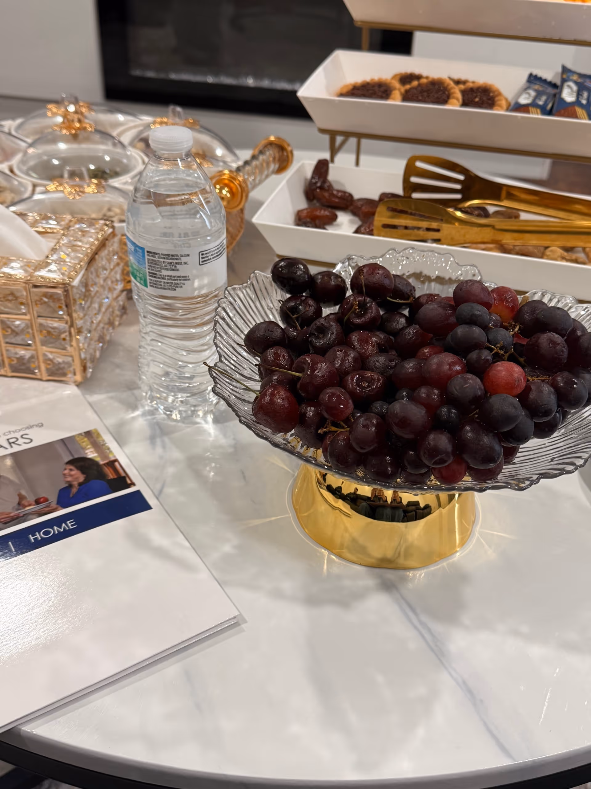 A crystal fruit pedestal with grapes sits on a marble table beside water bottles and pastries.