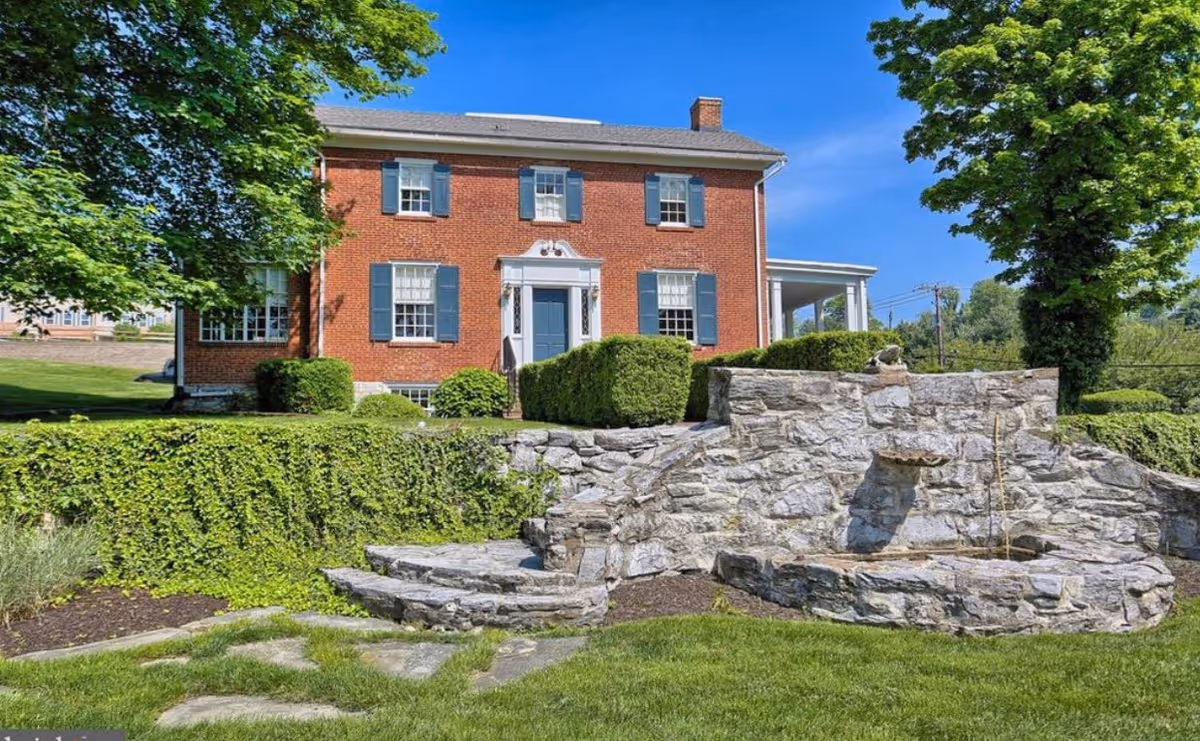Red brick senior living building with blue shutters sits behind stone wall and green landscaping.