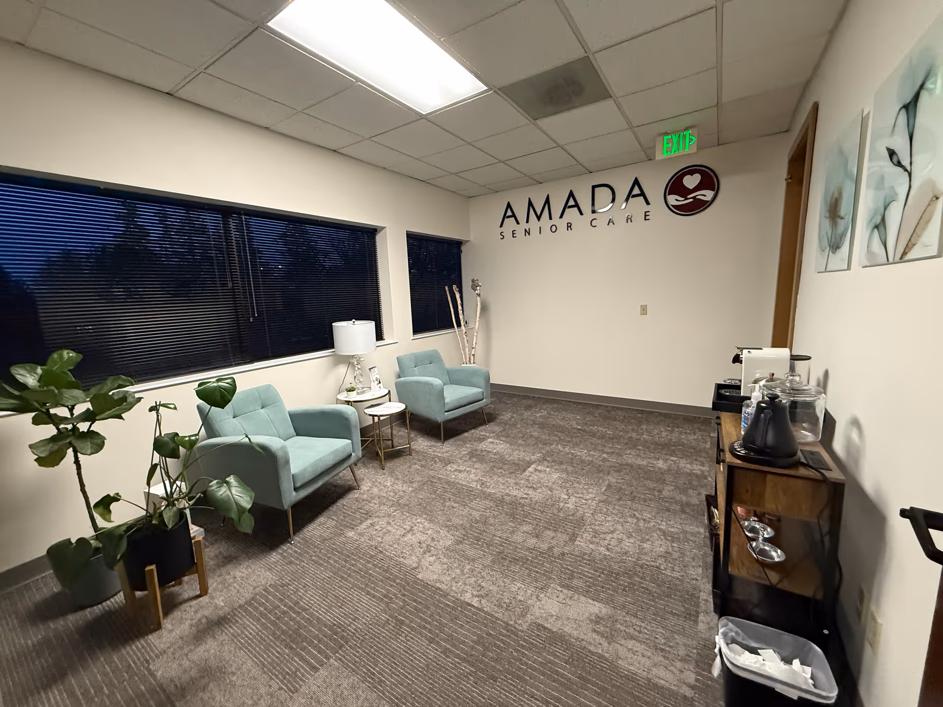 A quiet lobby with turquoise chairs, plants, and a coffee station beneath Amada Senior Care signage.