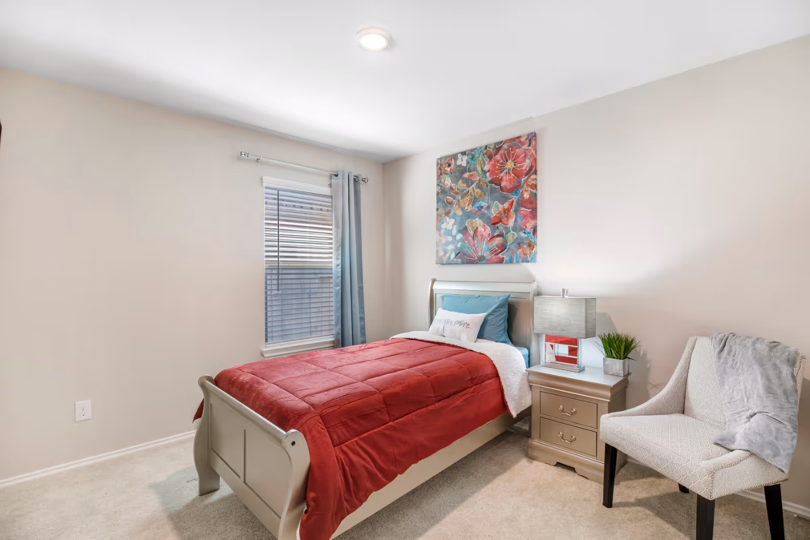 A neatly made bedroom with a single bed, nightstand, chair, and colorful wall art.