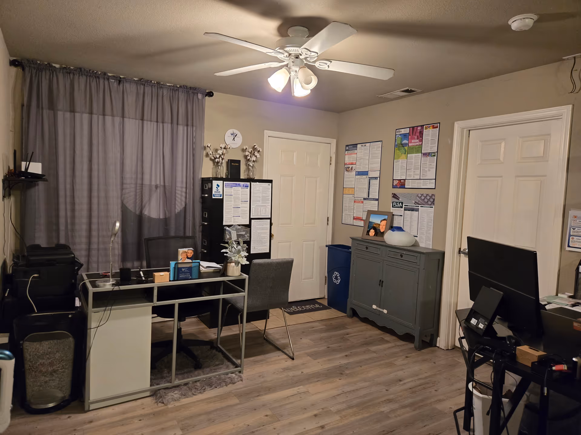 A small interior office space with desk, chairs, computer, bulletin boards, and ceiling fan.