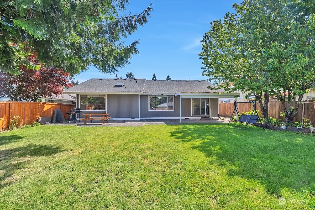 Single-story house with a spacious backyard, green lawn, trees, and outdoor seating.