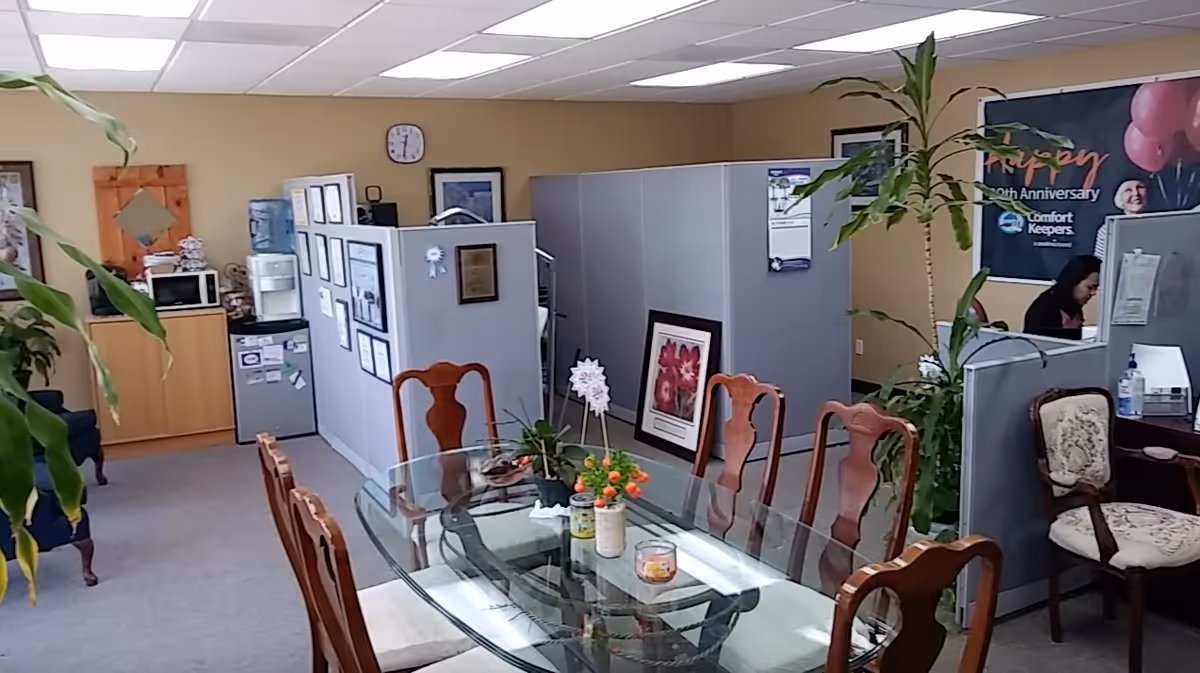 Office-style common area with a glass dining table, cubicles, plants, and a staff member.