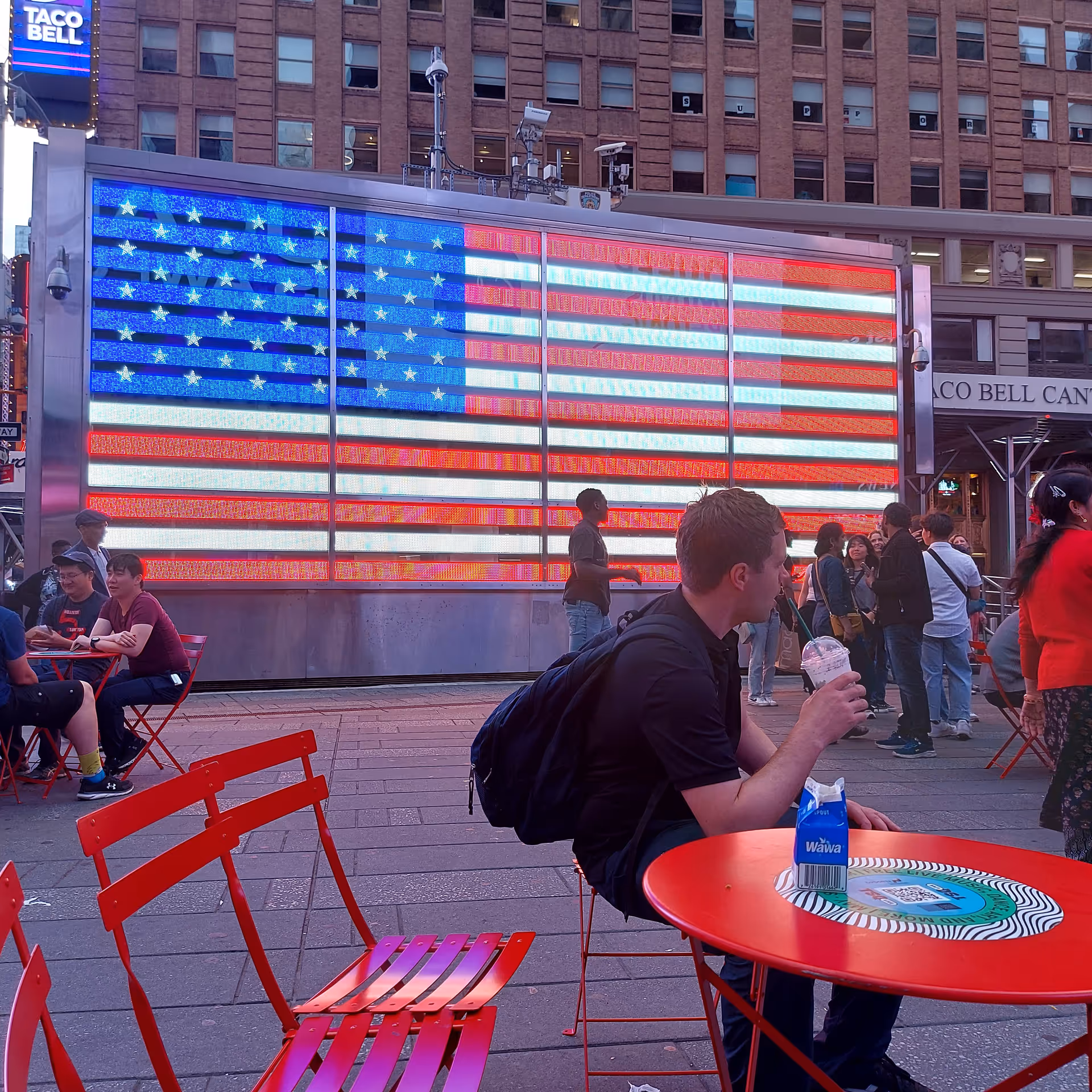 Public outdoor plaza with red tables and chairs, a giant illuminated American flag display.