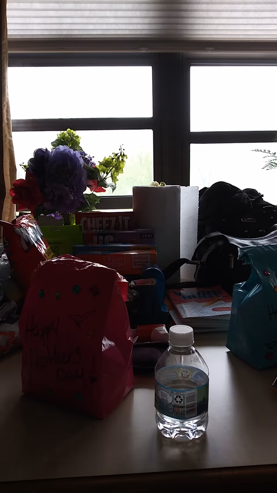 Cluttered desk with gift bags, a bouquet, and a water bottle by a window.