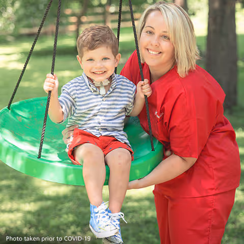 A caregiver in red scrubs pushes a smiling child on a green outdoor swing.