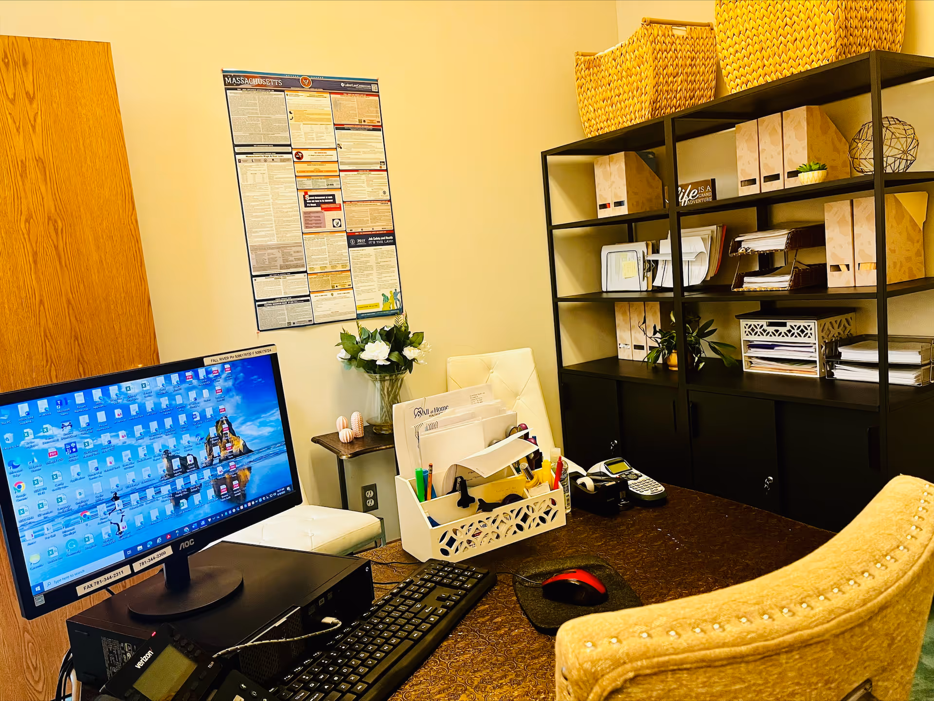 Office workspace with computer, filing shelves, and supplies in a warm, organized room.