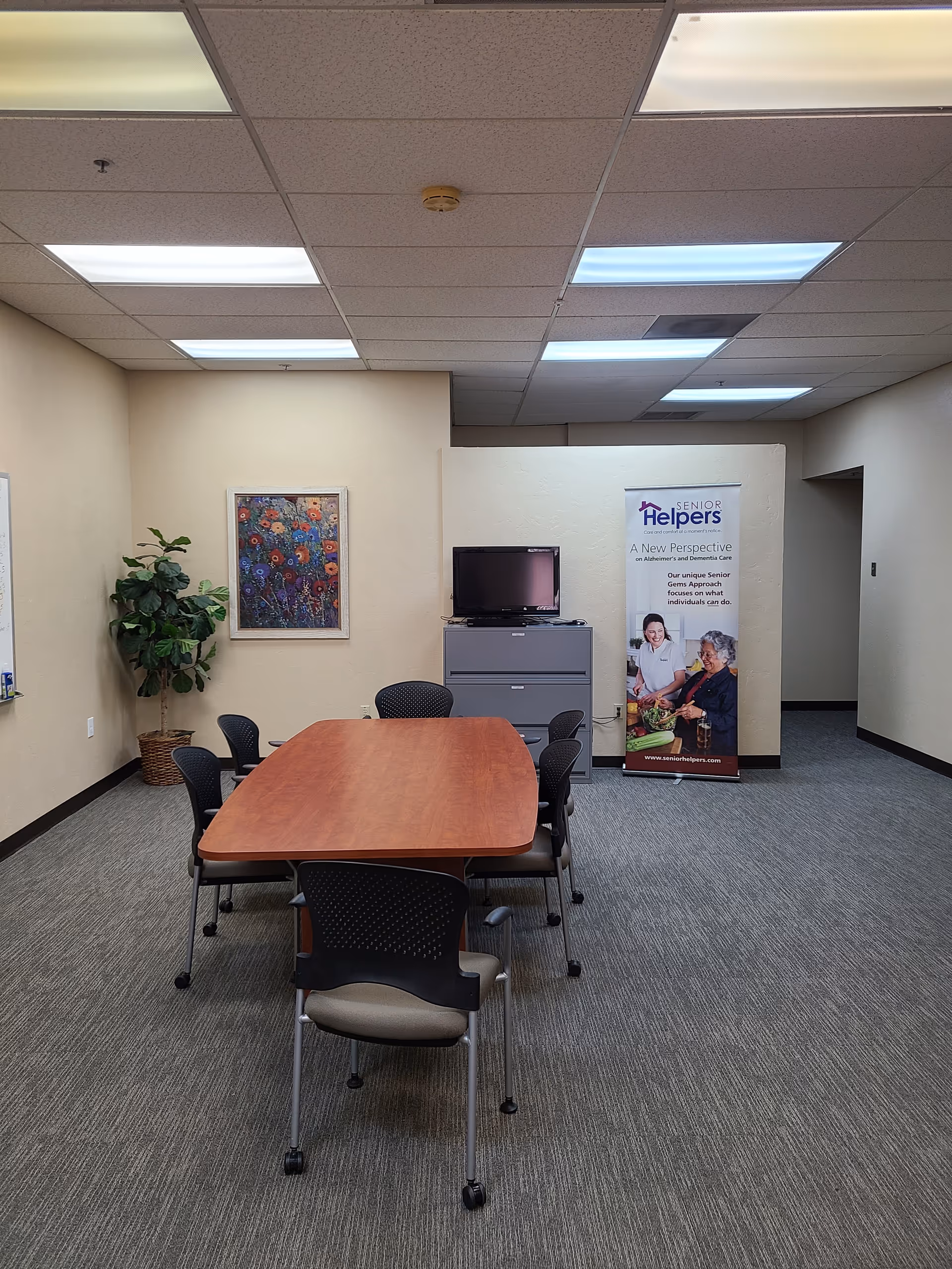 Interior meeting room at Senior Helpers with table, chairs, banner, TV, and artwork.