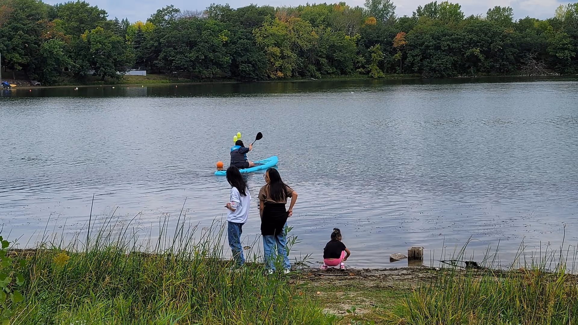 Several people at a lakeside, with a kayaker on the water and trees behind.