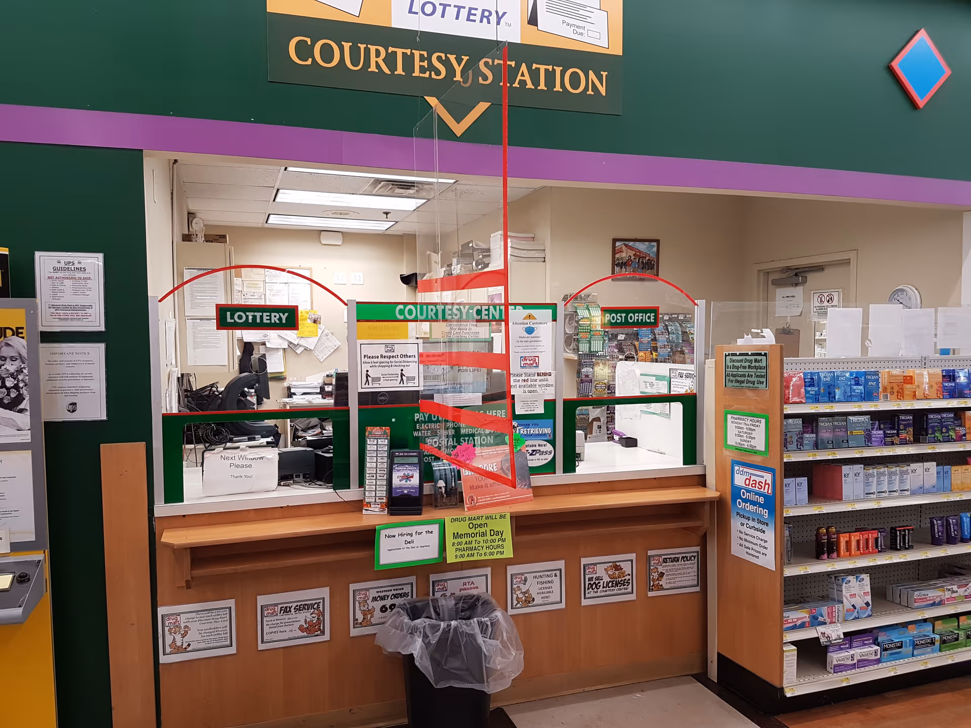 Interior view of a pharmacy counter labeled courtesy station with lottery and post office signs.