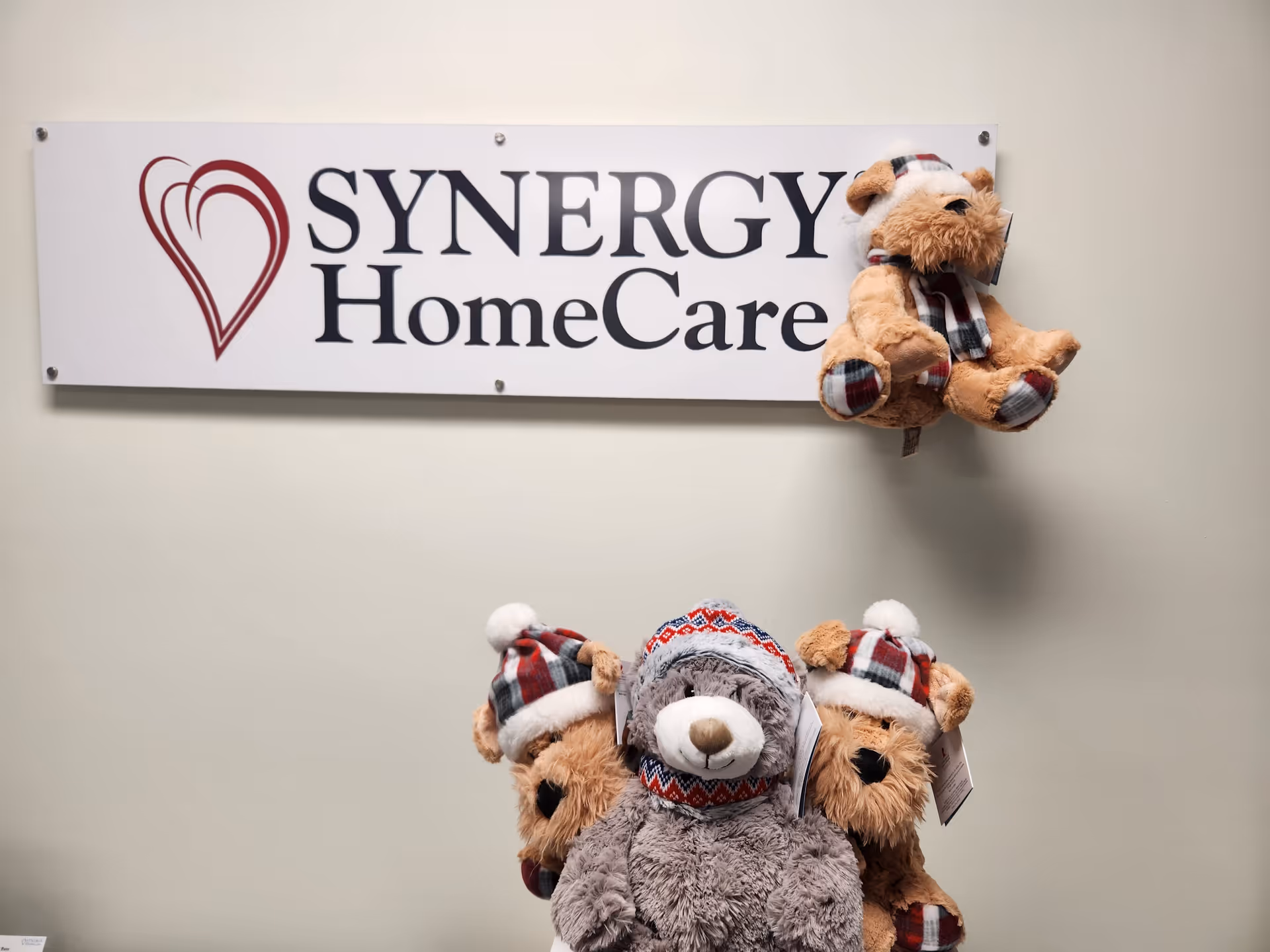 Wall sign reading SYNERGY HomeCare with plush bears wearing winter hats beneath.
