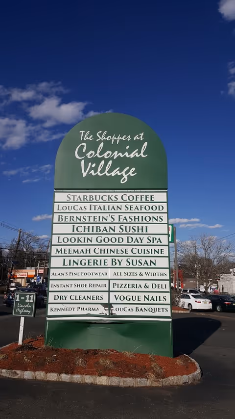 Green outdoor signboard displaying multiple store names in a parking lot under blue sky.