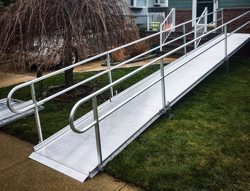 Metal outdoor wheelchair ramp with handrails beside a grassy lawn and facility entrance.