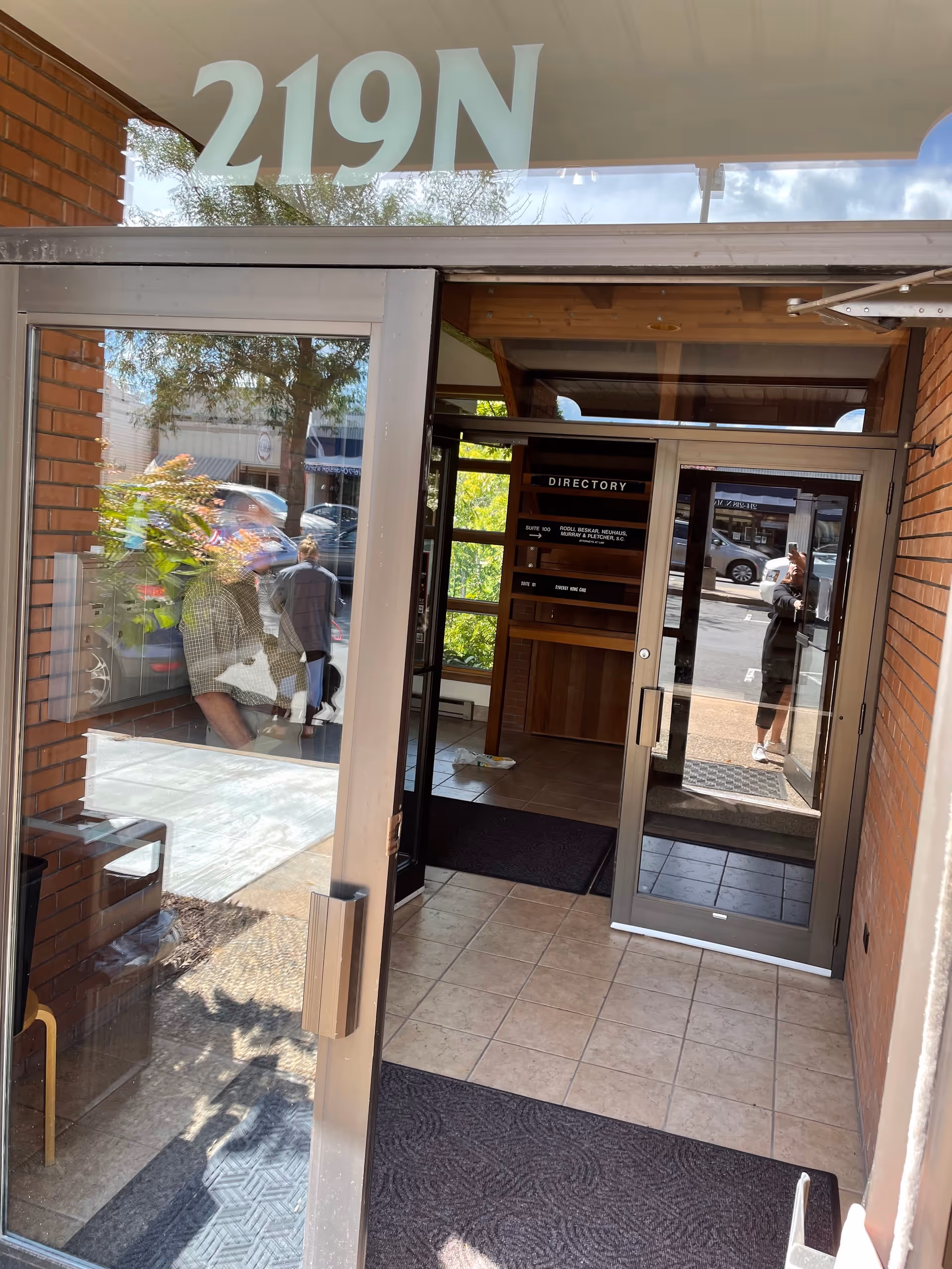 Glass-front entrance with a directory sign inside, brick exterior, and reflected pedestrians.