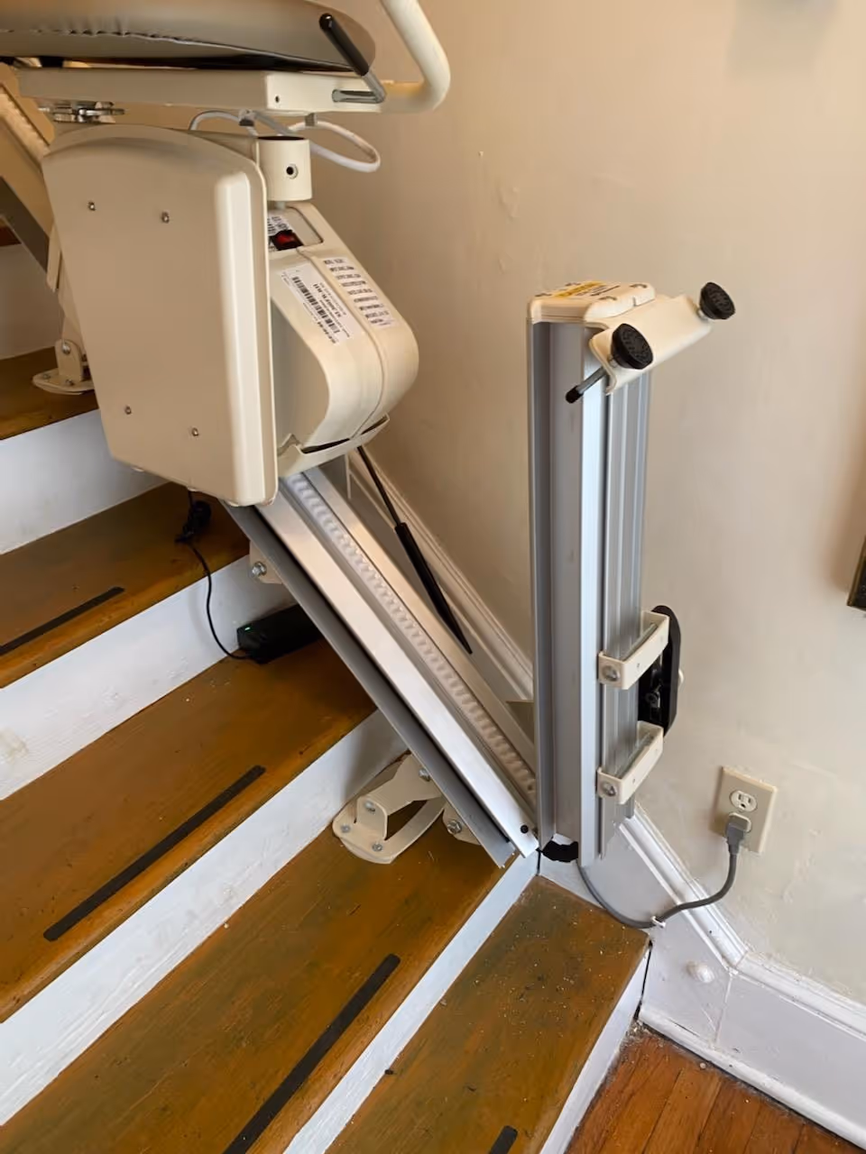 Indoor stair lift mounted along wooden stairs with power cord plugged into wall outlet.