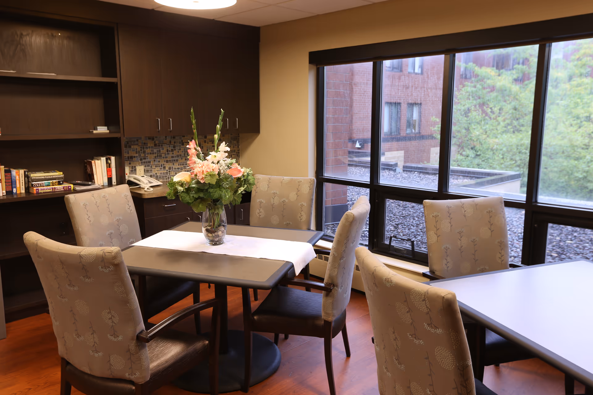 Interior dining area with tables, upholstered chairs, a flower centerpiece, and large windows.