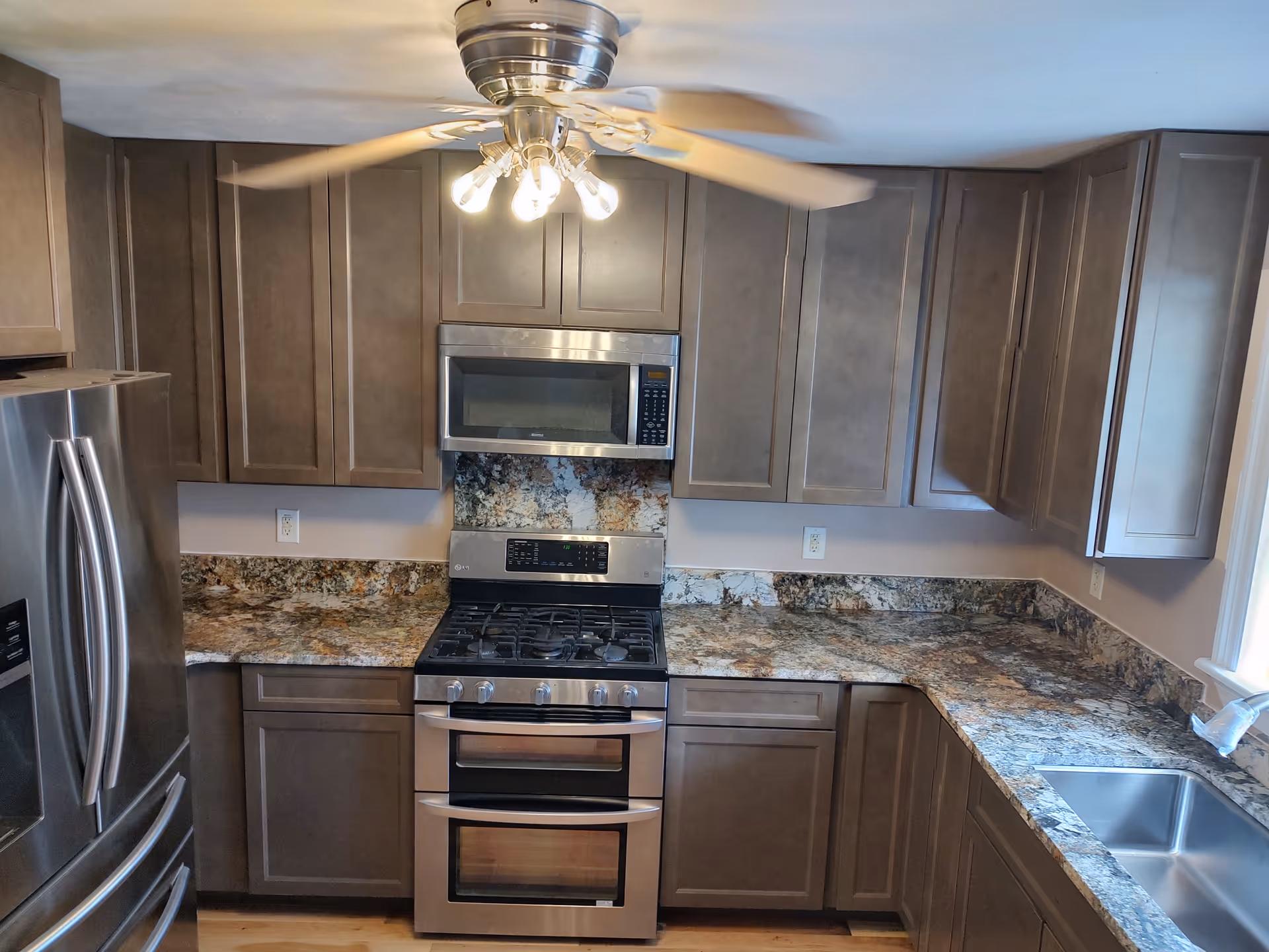 Modern kitchen with granite countertops, brown cabinets, stainless steel appliances, and a ceiling fan.
