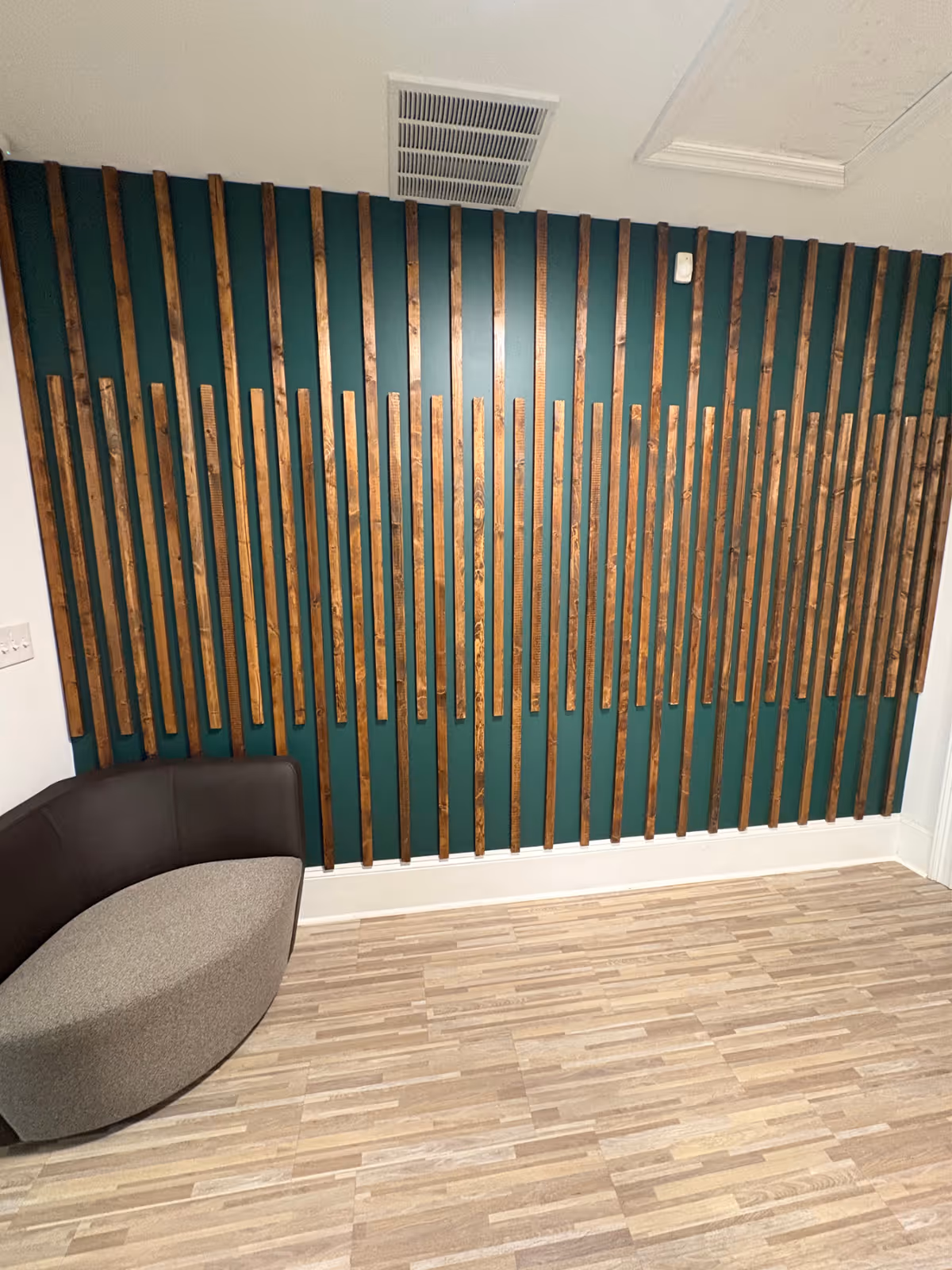Teal wall with vertical wooden slats and a curved lounge seat, laminate floor.