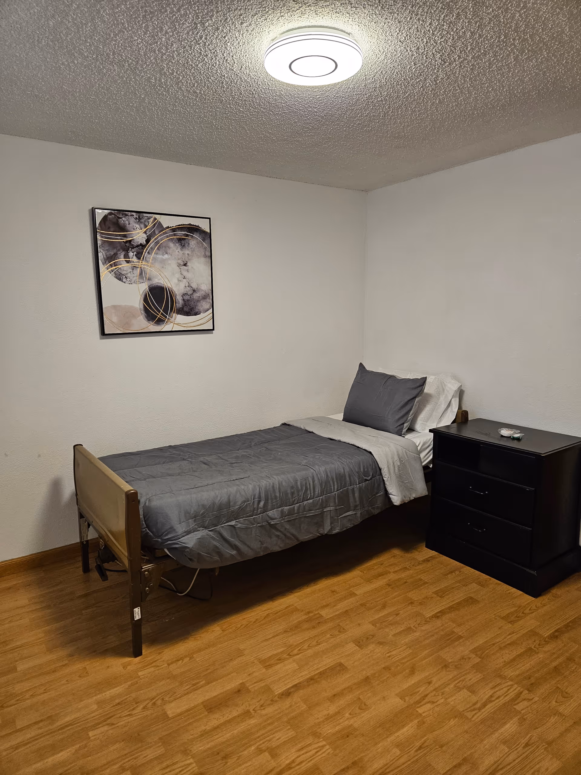 A small bedroom with a single bed, gray bedding, and a dark nightstand.