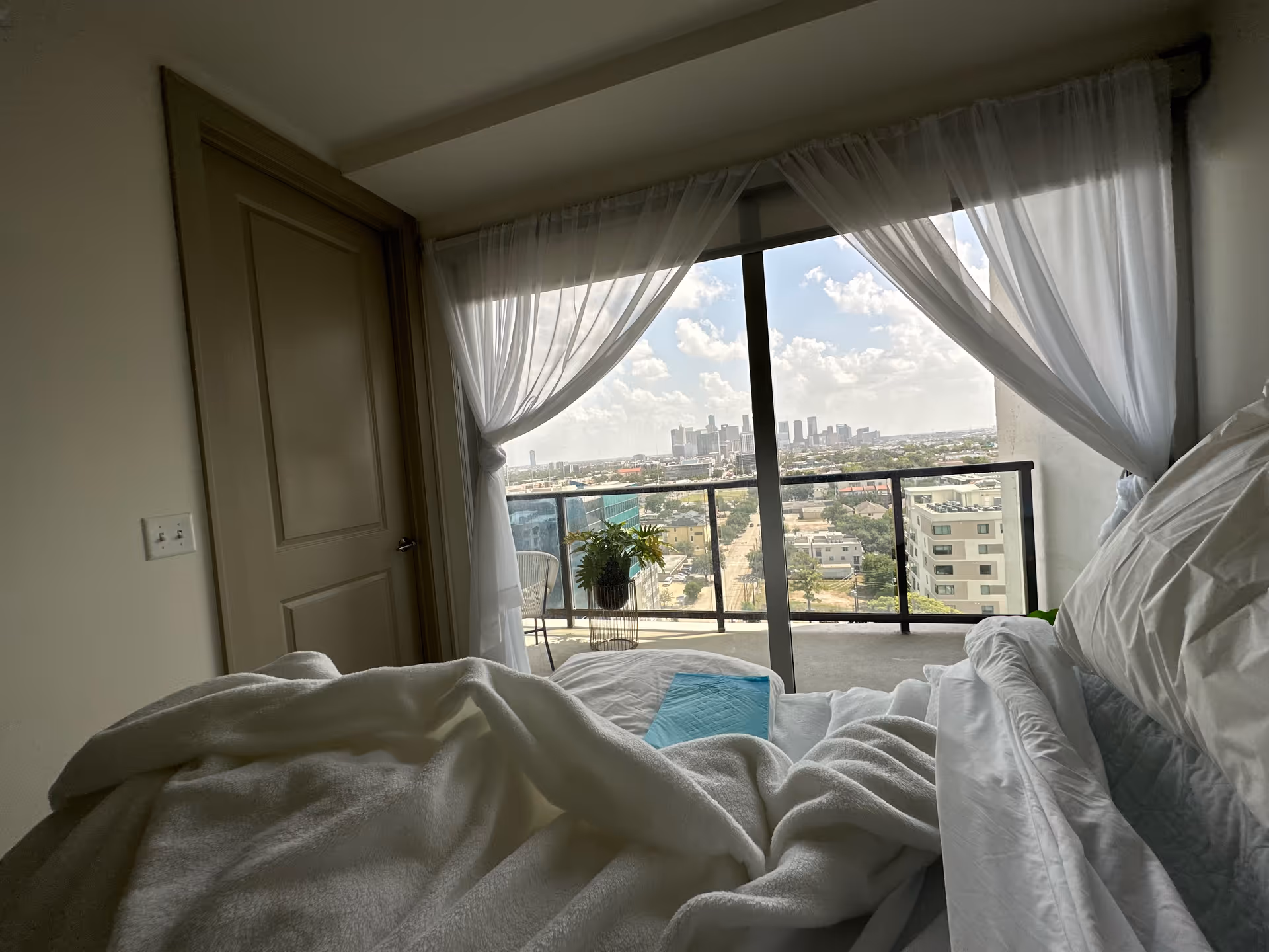 A bedroom with white bedding, sheer curtains, and a balcony overlooking a cityscape.