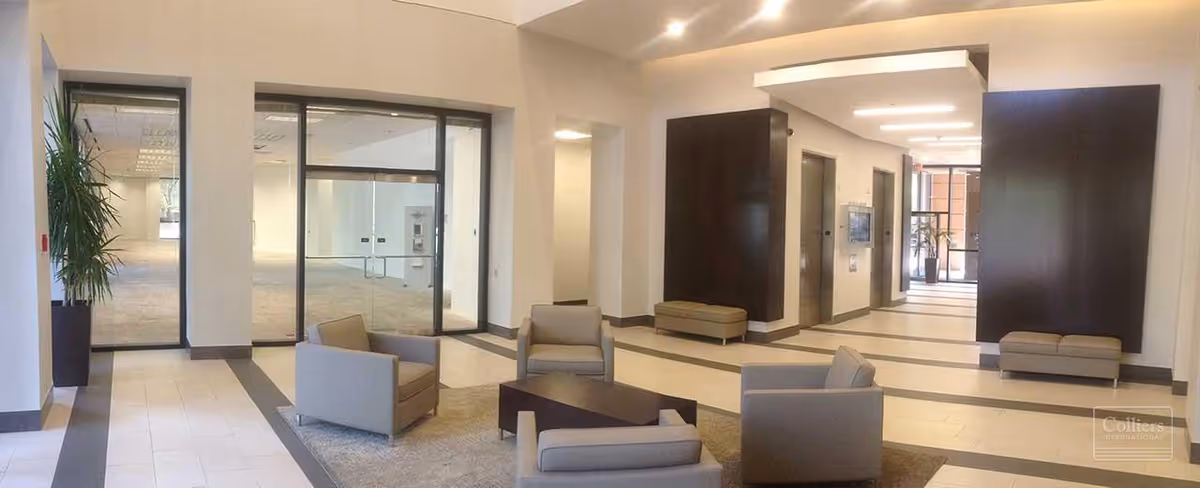 Wide bright lobby with gray sofas, glass doors, and potted plants.