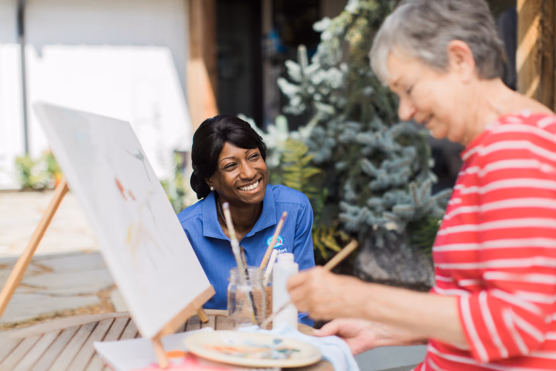 A smiling caregiver watches an elderly resident paint at an outdoor art table.