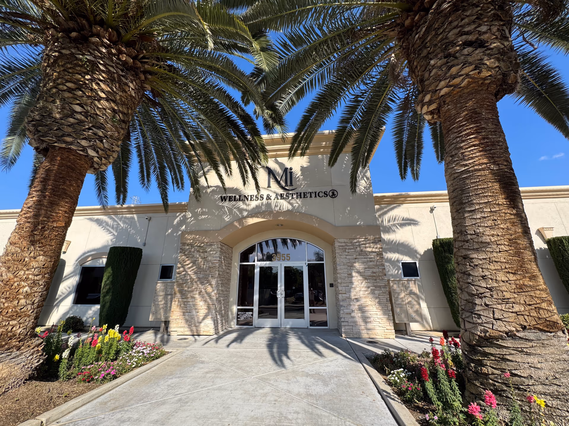The front entrance of a beige building is framed by palm trees and colorful flowers.