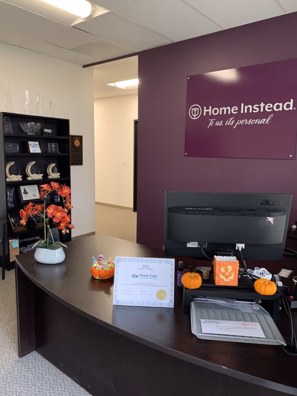 Lobby reception area with computer, purple Home Instead sign, awards shelf, and pumpkins.