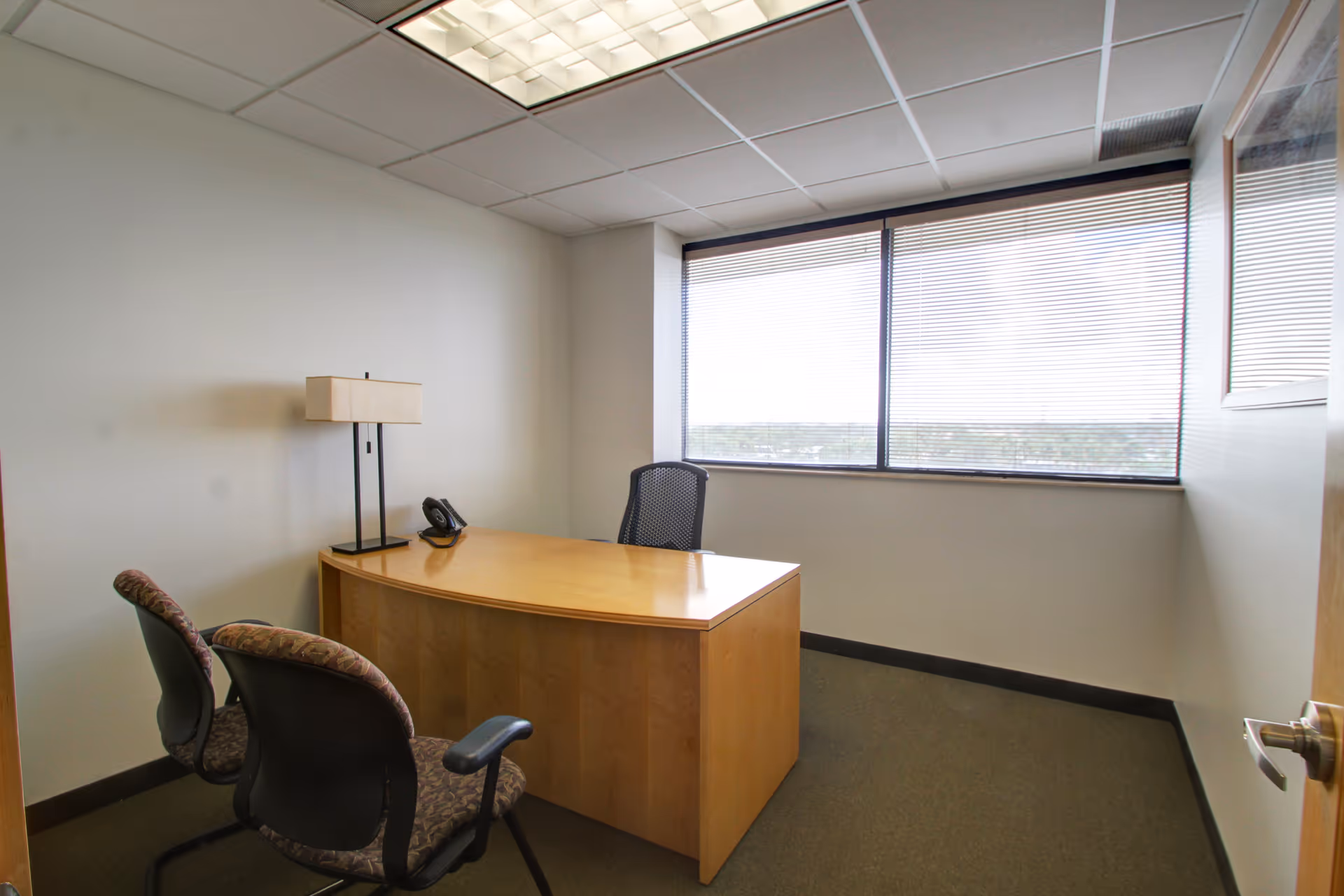 A small office room with a wooden desk, chairs, lamp, and large window.