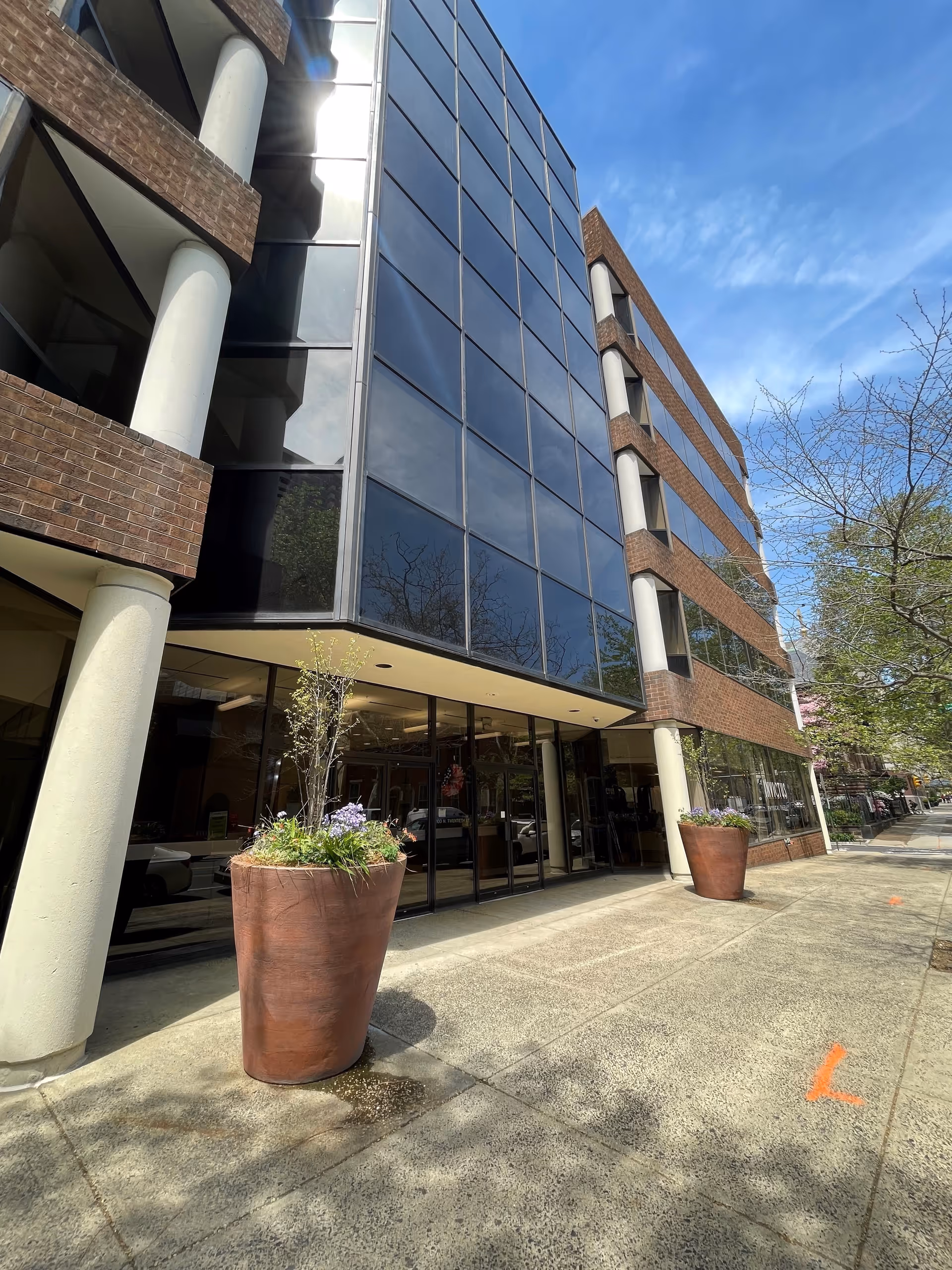 Modern glass-brick building exterior with large windows, planters, and a sidewalk.