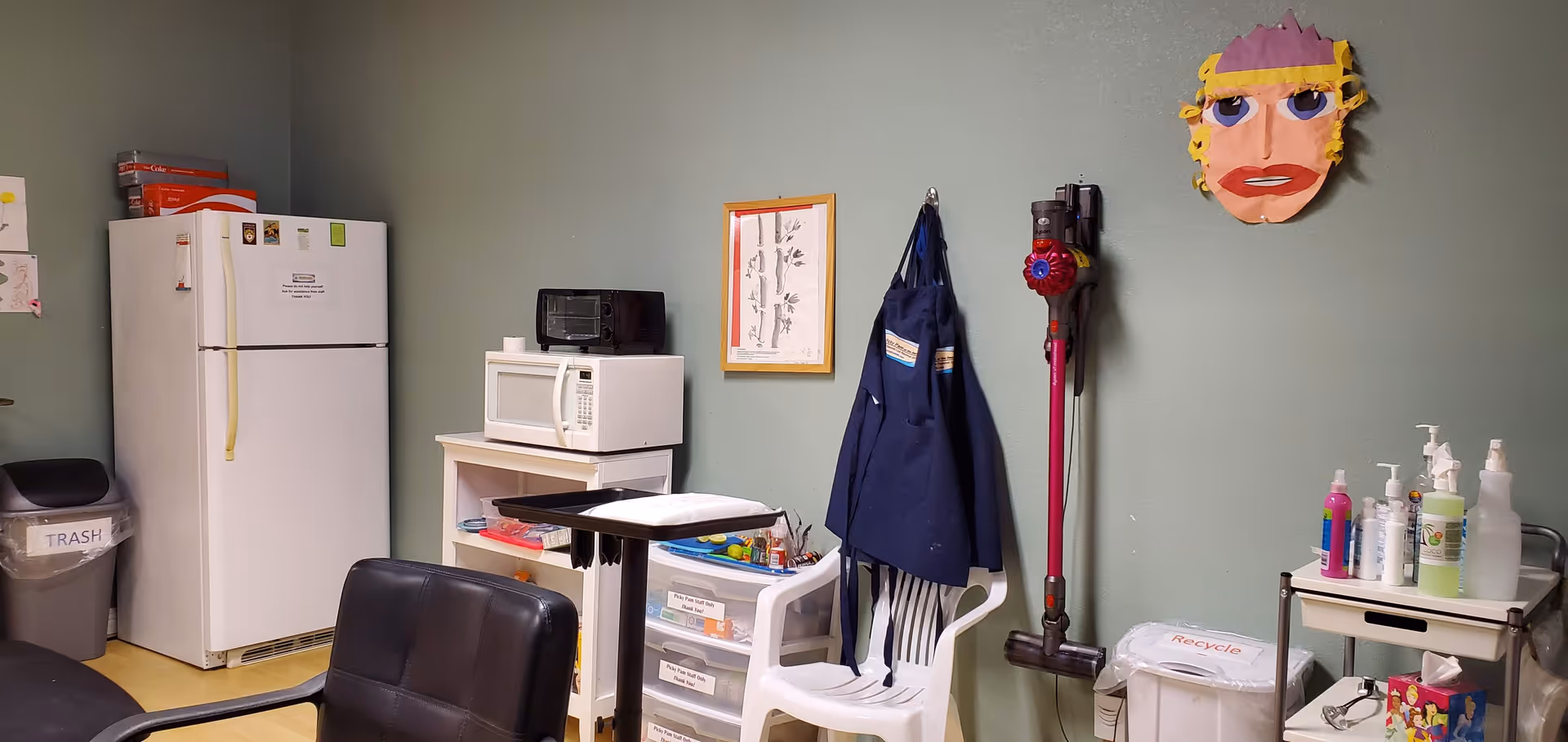 Compact kitchenette with fridge, microwave, stacked appliances, trash can, chair, coat, and cleaning supplies.