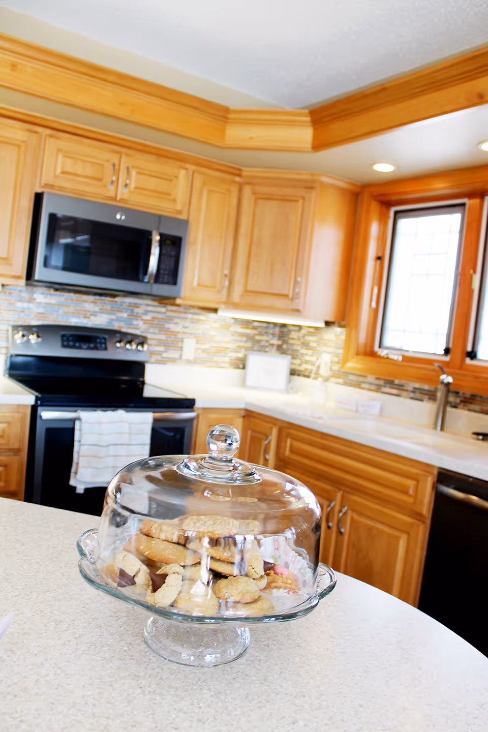 Warm wooden kitchen featuring an island, glass cake stand with cookies, and stainless appliances.