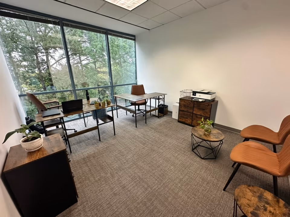 Office-like meeting room with desks, chairs, printer, plants, and large windows overlooking trees.