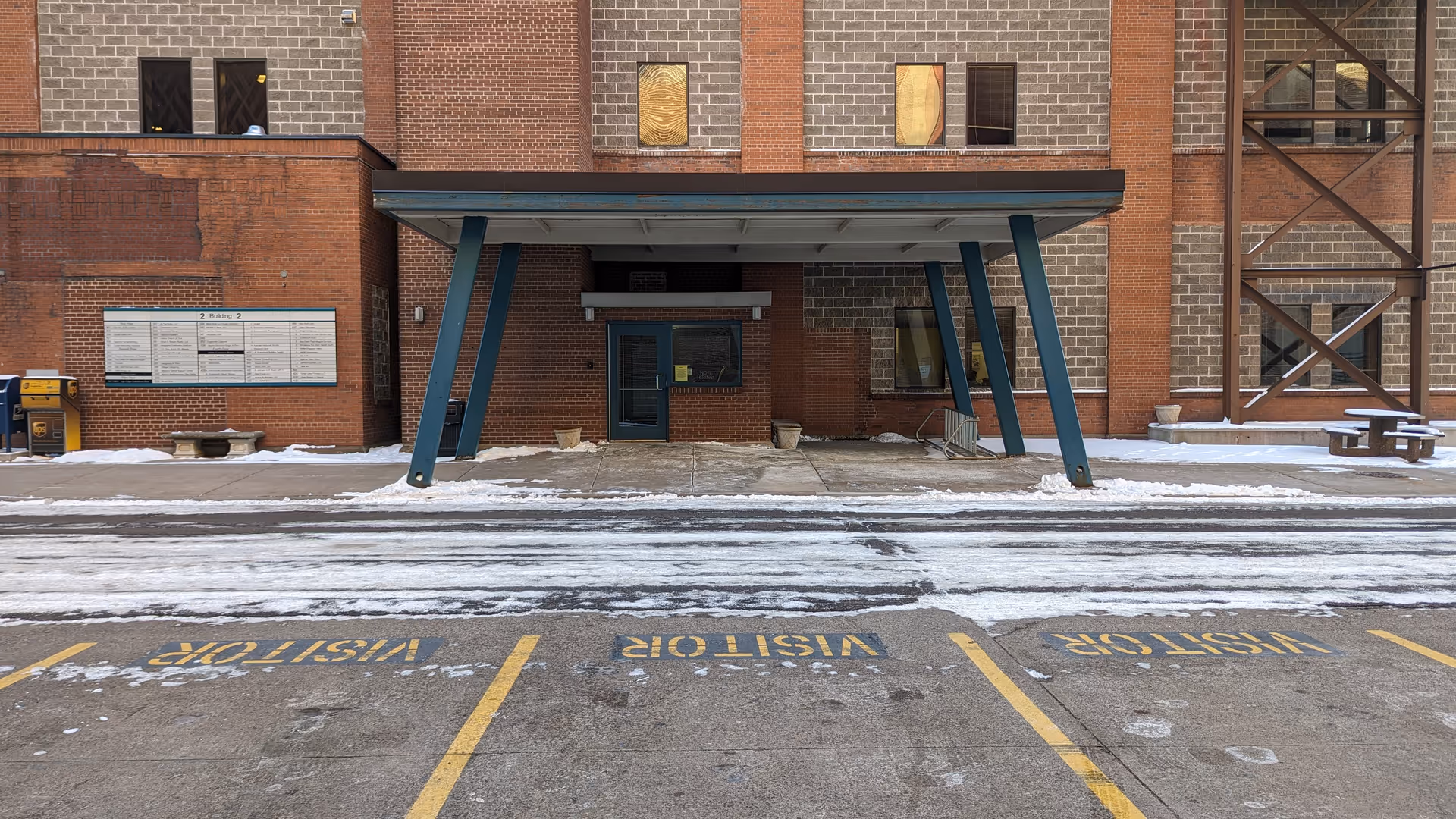 Brick senior living building entrance with blue angled canopy and snow-covered sidewalk.