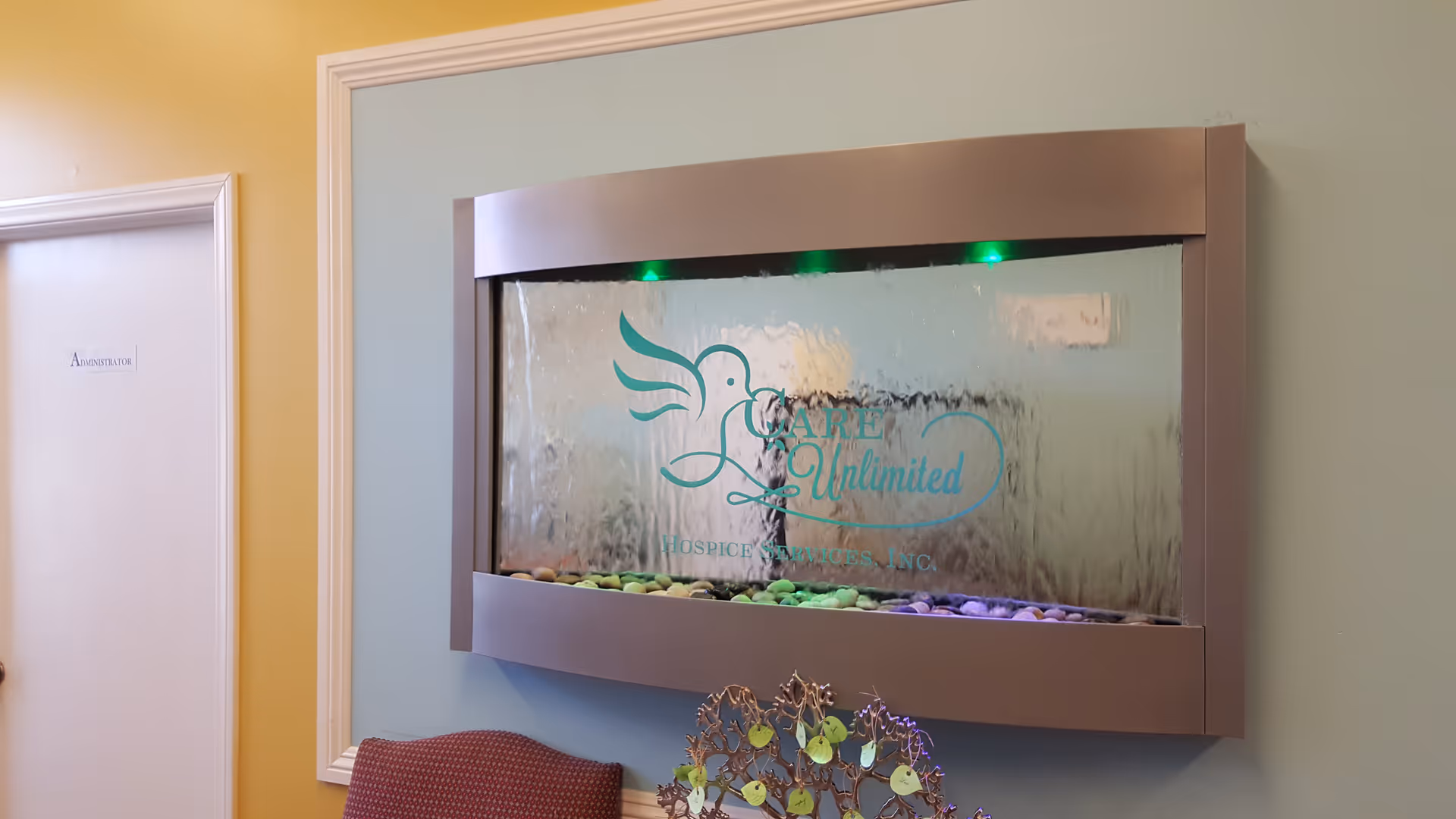 Wall-mounted glass water feature with pebbles and Care Unlimited logo inside a pastel interior